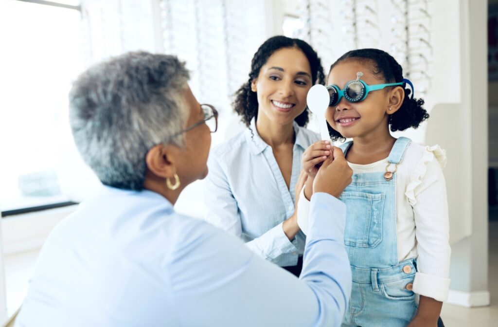 A child wears eye exam glasses and smiles as an optometrist guides her through a vision test.